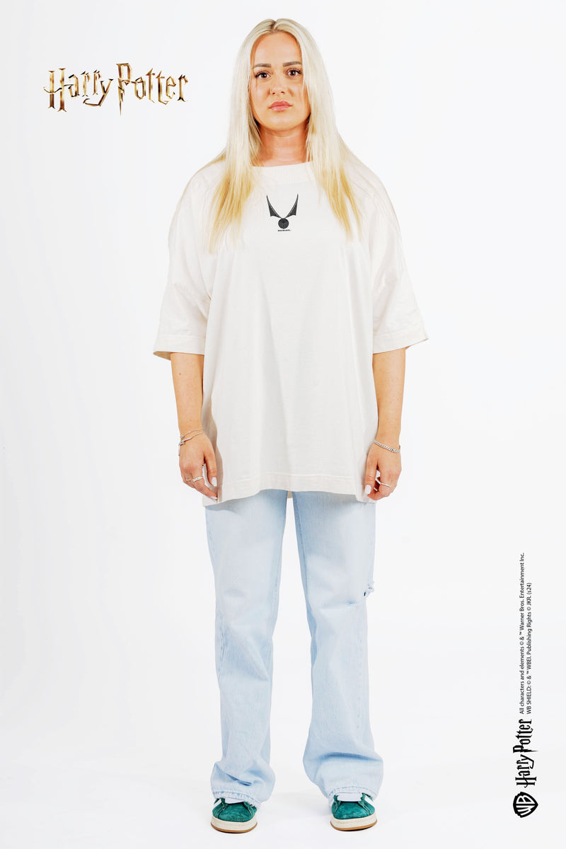 Frau mit blondem Haar trägt ein weißes Harry-Potter-T-Shirt mit einem Schnatz-Design und steht vor einem neutralen, weißen Hintergrund.